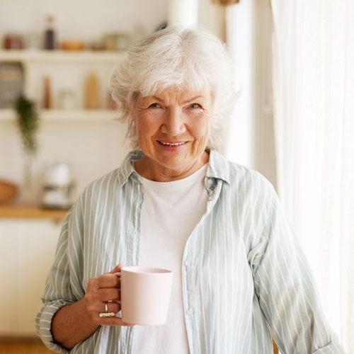 Bild einer fröhlichen, glücklichen europäischen Rentnerin, die sich zu Hause entspannt, Kräutertee trinkt, breit in die Kamera lächelt und vor einem Küchenhintergrund posiert